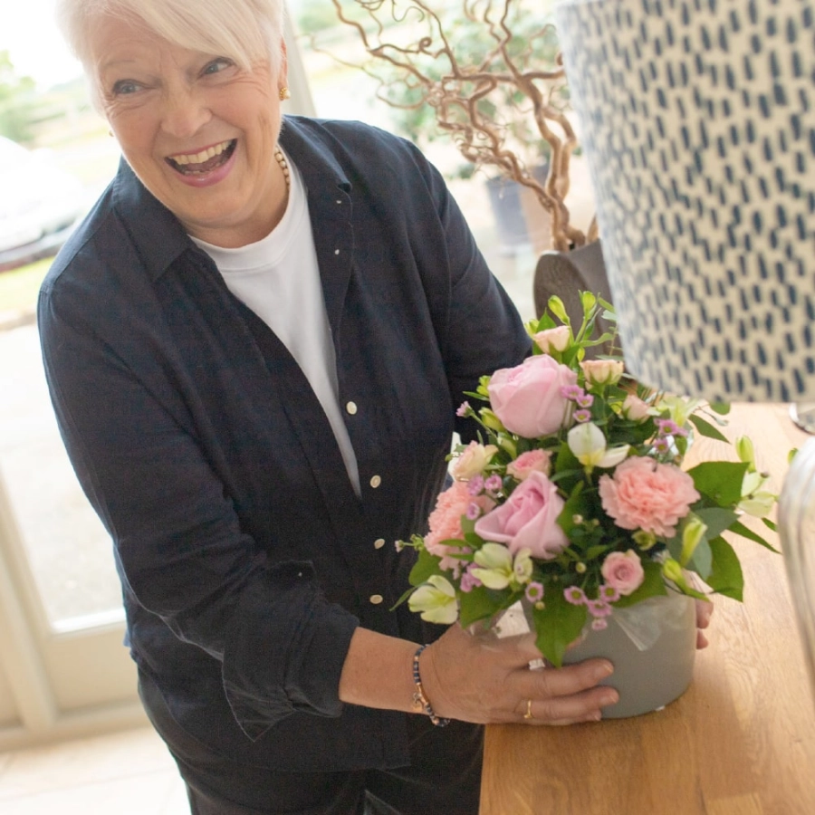 A smiling woman places a beautiful pink and green florist-choice arrangement on a wooden table near a bright window choice arrangement on a wooden table near a bright window