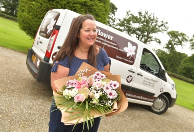 Woman florist with delivery van behind her holding a pink wrap of flowers she's delivering.