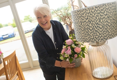 Older woman laughing and looking at someone whilst placing a pot of flowers on a sideboard.