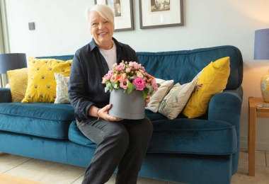 Elder woman wearing navy sitting on sofa holding a grey hatbox arrangement filled with pink and peach flowers